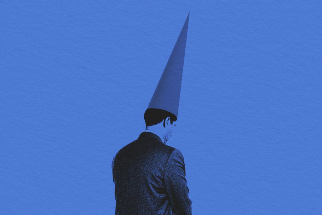 businessman sitting on a stool, with his back to the camera, wearing a dunce cap